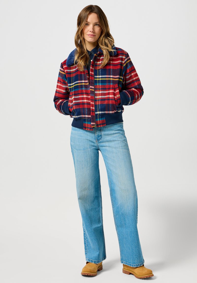 Czerwono-granatowa kurtka bomberka w kratę z futrzanym kołnierzem, ściągaczami przy mankietach i dolnej krawędzi, zestawiona z jasnoniebieskimi dżinsami w stylu dzwonów i brązowymi botkami za kostkę.