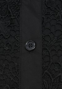 Čierna látka s komplikovanou čipkovanou aplikáciou na ľavej strane. Veľké čierne gombík je umiestnený vpravo vedľa hladkej textúrovanej časti.