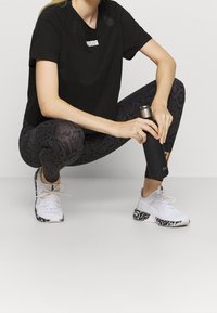 Svart tränings-T-shirt med logga, mönstrade svarta leggings och vita sneakers med svarta detaljer; person som håller en svart vattenflaska.