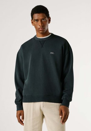 Joven con sudadera negra de PJL y pantalones de pana beige, de pie frente a un fondo claro y liso con una expresión neutral.