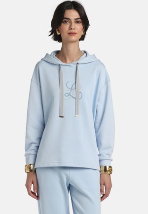 Kvinde iført lyseblå hættetrøje med monogramdetalje, matchende bukser, guldbelagte øreringe og guldcuff-armbånd, stående foran en ensfarvet baggrund.