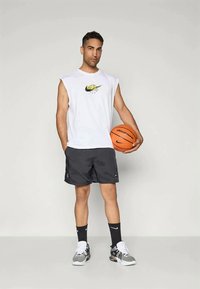 Valkoinen hihaton paita, jossa on keltainen ja musta graafinen logo, mustat urheilushortsit, valkoiset ja mustat lenkkarit, pitäen oranssia koripalloa.