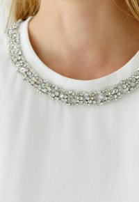 Weißes Oberteil mit rundem Ausschnitt, verziert mit einer dekorativen silbernen Perlen- und Kristallverzierung, die eine glatte Textur zeigt.