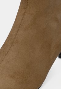 Bottine en daim marron avec une texture lisse, une forme effilée, et un petit talon noir. Des coutures décoratives longent le côté.