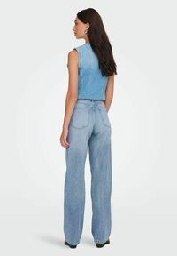 Femme aux longs cheveux foncés, portant un gilet en jean sans manches et un jean taille haute à jambes larges, debout de profil contre un fond blanc.