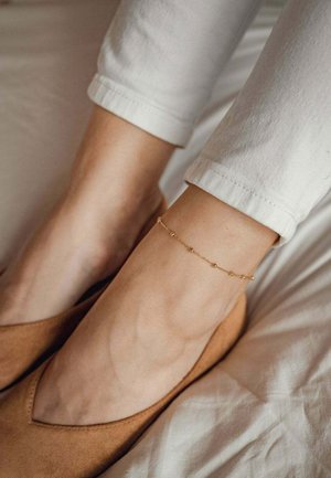 Goldenes Knöchelbändchen mit kleinen runden Perlen, das auf einem nackten Knöchel liegt, kombiniert mit beigen Wildlederschuhen. Weiße, hochgekrempelte Hose sichtbar.