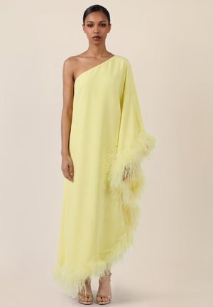 Frau in einem bodenlangen, gelben Ein-Schulter-Kleid mit Federbesatz entlang des Ärmels und Saums, stehend vor einem schlichten Hintergrund.