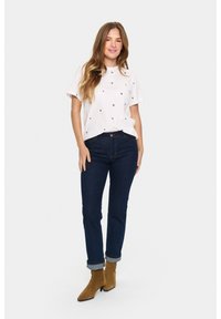T-shirt blanc avec un petit motif de cœurs navy, associé à un jean bleu foncé avec des manches retroussées. Bottines beiges à bout pointu.