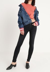 Chaqueta de mezclilla con puños a rayas rojas y blancas, camiseta rosa corta, jeans ajustados negros y mules negros. Pose relajada contra un fondo simple.