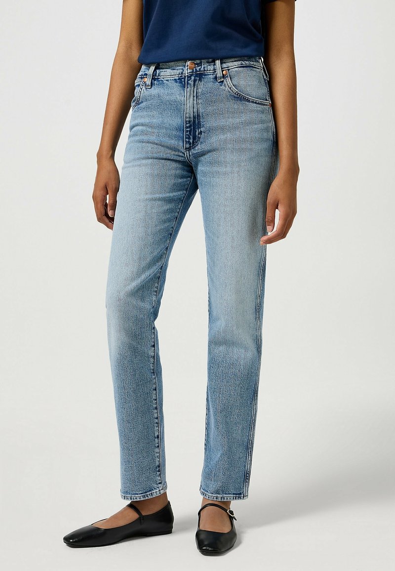 Jeans de mezclilla azul claro con corte de pierna recta, diseño de cinco bolsillos y un sutil desgaste. Combinados con zapatos planos negros y una blusa azul marino.