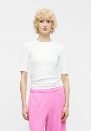 Szőke göndör hajú személy fehér rövid ujjú felsőt és élénk rózsaszín nadrágot visel, sima világos háttér előtt áll.