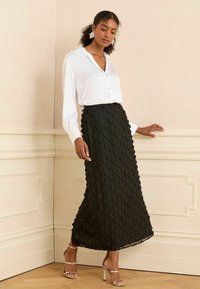 Jupe midi à motif floral noir avec un design texturé, associée à une chemise blanche à boutons. Le mannequin porte des talons argentés à lanières.