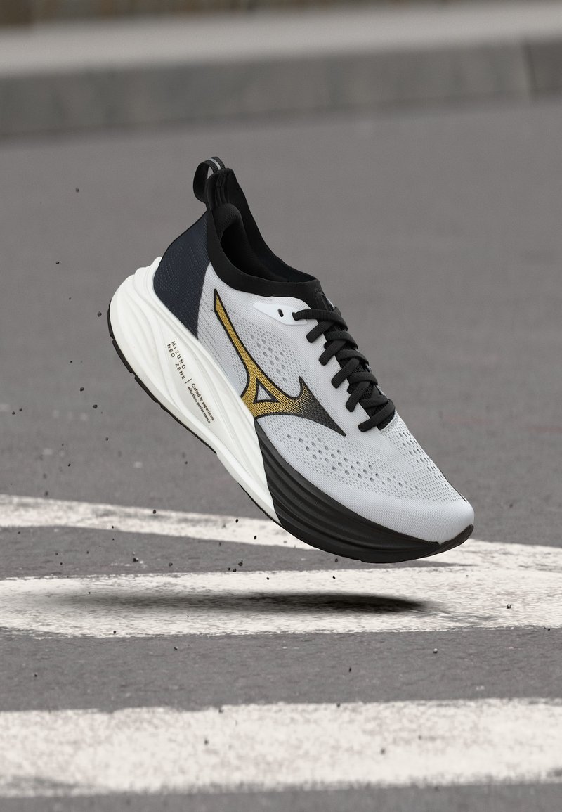 Γκρι και μαύρο αθλητικό παπούτσι με χρυσό λογότυπο, στον αέρα πάνω από άσφαλτο με λευκές λωρίδες δρόμου, με σωματίδια βρωμιάς γύρω από τη σόλα.