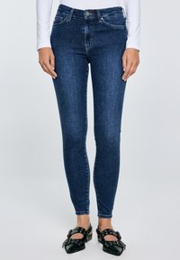 Žena u tamnoplavim uskim trapericama, bijeloj majici s dugim rukavima i crnim špicastim cipelama s ukrasnim kopčama i rupicama.