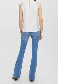 Top bianco senza maniche con dettagli in pizzo sulle spalle e un'apertura a chiave sul retro, abbinato a jeans svasati di un blu chiaro. Colore uniforme, texture liscia.