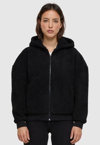 Black, hooded jacket made of textured fleece material. Features a zip closure and elastic cuffs. Casual, oversized fit with a soft exterior.
