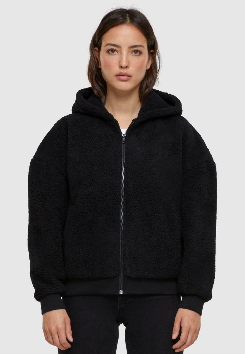 Black, hooded jacket made of textured fleece material. Features a zip closure and elastic cuffs. Casual, oversized fit with a soft exterior.