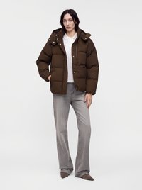 Femme portant une doudoune marron par-dessus une chemise blanche, un jean gris à jambes larges, et des chaussures marron à bouts pointus, debout devant un fond uni.