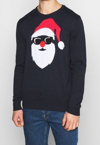 Mörkblå stickad tröja med en grafik av Santas ansikte, som har en röd mössa, vit skägg, svarta solglasögon och en röd näsa. Ribbad mudd och nederkant.