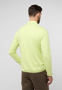 Helles grünes Langarmhemd mit Kragen, glatter Textur, elastischem Saum und taillierten Ärmeln. Trägt dazu eine dunkelbraune Hose.