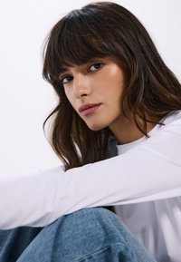 Primo piano di una persona con capelli lunghi e scuri e frangia, che indossa una maglietta bianca a maniche lunghe e jeans blu, con pelle morbida e trucco naturale.
