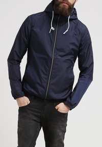 Marinblå vindjacka med huva, dragkedja, elastiska ärmslut och dragsko-accenter. Slätt, lättviktigt material.