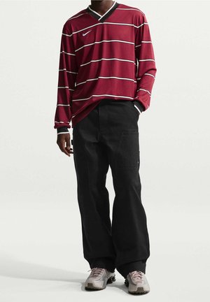 Personne portant une chemise rayée bordeaux à manches longues, un pantalon noir à jambe large et des baskets blanches, debout devant un fond uni.