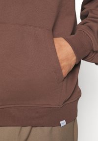 Brauner Pullover mit Vordertasche, gerippten Bündchen und Saum, mit einem Etikett unten rechts. Glatter, strukturierter Stoff.