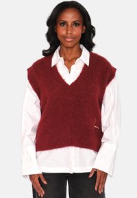 Burgunderfarget strikket vest med V-hals, lagt over en hvit skjorte med krage. Har brede armåpninger og en myk, lodden tekstur.