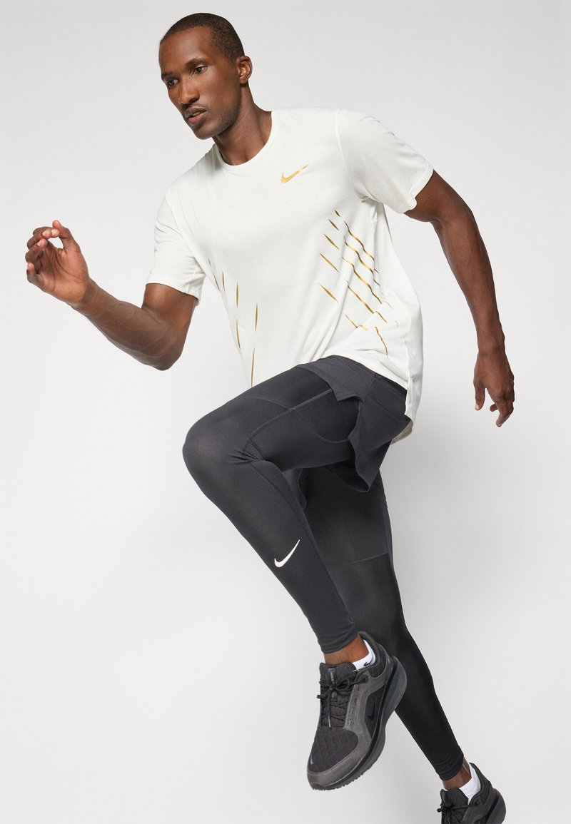 Męska odzież sportowa składa się z jasnoszarej koszulki z krótkim rękawem z złotymi detalami oraz czarnych legginsów z białym logo Nike, do których dobrano czarne buty sportowe.