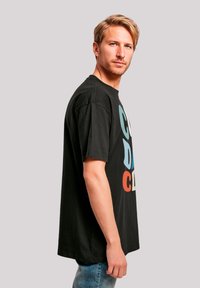 Black cotton T-shirt with large multicolored text design. Short sleeves, relaxed fit, and visible seam detailing. Paired with light denim jeans.