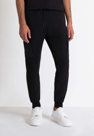 Joggers negros con un diseño ajustado, cintura elástica y puños de canalé. Presentan dos bolsillos laterales y una tela suave y texturizada. Combinados con zapatillas blancas.