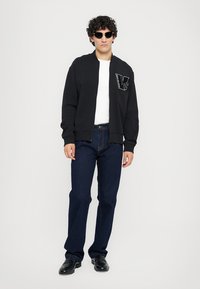 Črna bomber jakna z zadrgo in vezenim logotipom, bela majica, temno modri kavbojski hlače in črne čevlje; model nosi sončna očala.
