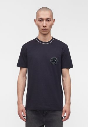 Homme au crâne rasé portant un t-shirt noir à manches courtes avec un logo rond sur la poitrine gauche, debout devant un fond blanc uni.