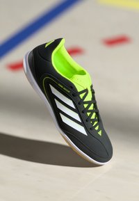 Scarpa da calcetto nera con accenti giallo brillante, caratterizzata da tre strisce bianche, una parte superiore testurizzata e una suola in gomma.