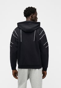 Άνδρας με την πλάτη, φοράει μαύρο φούτερ με λευκές ρίγες στα μανίκια και στην πλάτη, σε συνδυασμό με ανοιχτό γκρι φόρμα, σε απλό φόντο.