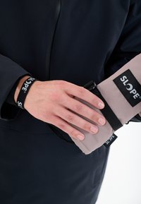 Manchette en tissu gris léger avec une sangle noire et un logo. Main dans un manteau noir tenant la manchette, mettant en valeur une texture lisse.