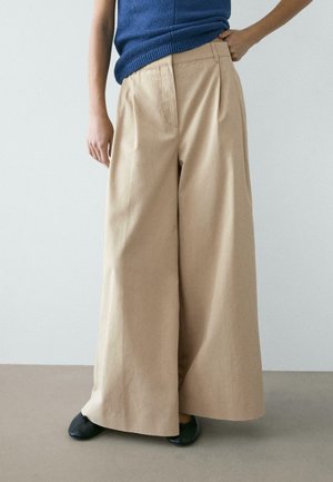 Persoon die beige wijduitlopende broek, blauw mouwloos topje en zwarte schoenen draagt, staat tegen een effen achtergrond.