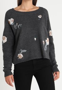 Ciemnoszary sweter dzianinowy z beżowymi i białymi kwiatowymi wzorami, noszony z czarnymi spodniami, z okrągłym dekoltem i długimi rękawami.