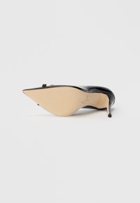 Μαύρη γυναικεία γόβα από λουστρίνι με μυτερή μύτη, λεπτό τακούνι και μπεζ σόλα. Διαθέτει μίνιμαλ σχεδιασμό και χρυσές λεπτομέρειες.