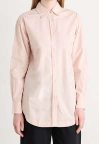 Person klädd i en ljusrosa långärmad skjorta med knappar och spetsig krage, samt svarta byxor, stående mot en enkel vit bakgrund.