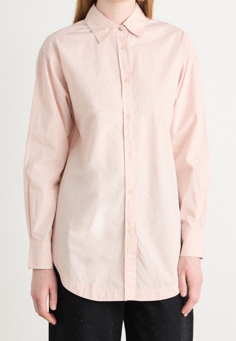 Person klädd i en ljusrosa långärmad skjorta med knappar och spetsig krage, samt svarta byxor, stående mot en enkel vit bakgrund.