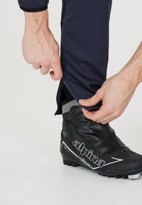 Svart längdskidboots med vit logotyp; hand som drar en dragkedja på marinblå byxor; byxorna har ett meshinlägg vid ankeln.