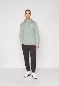 Light green hoodie with front pocket and logo, paired with dark gray sweatpants. Model wears white sneakers with black and red accents.