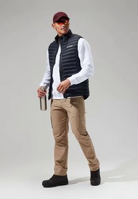Zwarte donzen bodywarmer over een wit shirt met lange mouwen, kaki cargobroek, zwarte laarzen en een bordeauxrode pet. Houdt een zilveren waterfles vast.