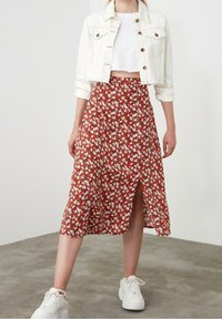 Jupe florale rouge avec une fente à l'avant, fabriquée en tissu léger. Le modèle porte un top court blanc et une veste en denim clair. Baskets blanches.