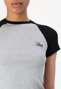 Sivý rebrovaný krátky rukáv s čiernymi raglánovými rukávmi, ktorý má na hrudi malú vyšitú značku "Guess Jeans".