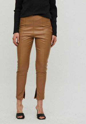 Pantalons en simili cuir beige avec une taille haute, une silhouette ajustée et des fentes sur les poignets. Associés à des talons ouverts noirs et un haut noir.