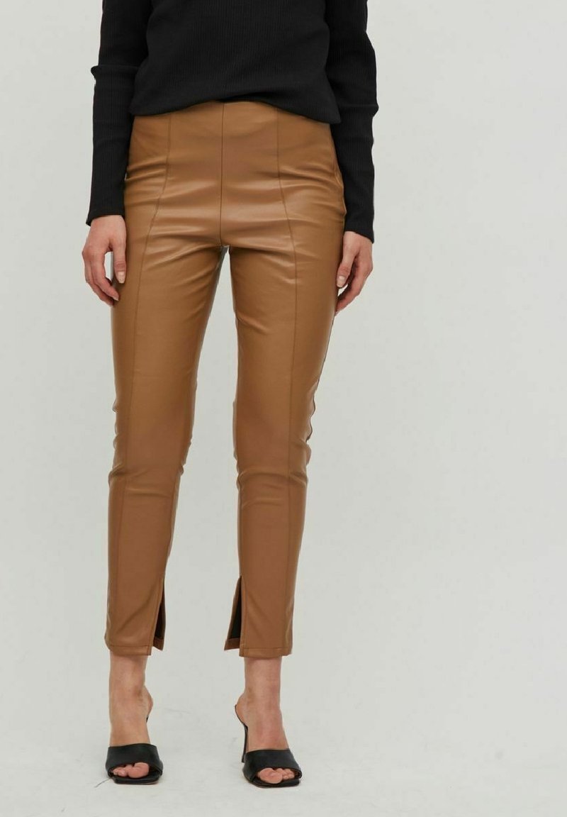 Pantalons en simili cuir beige avec une taille haute, une silhouette ajustée et des fentes sur les poignets. Associés à des talons ouverts noirs et un haut noir.