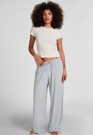 Vrouw met krullend haar, gekleed in een wit kort mouw crop top en losse lichtblauw gestreepte broek, staand op blote voeten tegen een effen achtergrond.